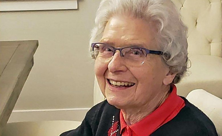Wilma Foubert, smiling, in blue glasses, a black cardigan, and a red collar